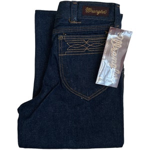 May include: A pair of dark blue denim jeans with a brown leather Wrangler label on the back waistband. The jeans have a boot cut flare and a brown embroidered design on the back pocket. The jeans have a tag that says "Wrangler" and "Boot Flare".