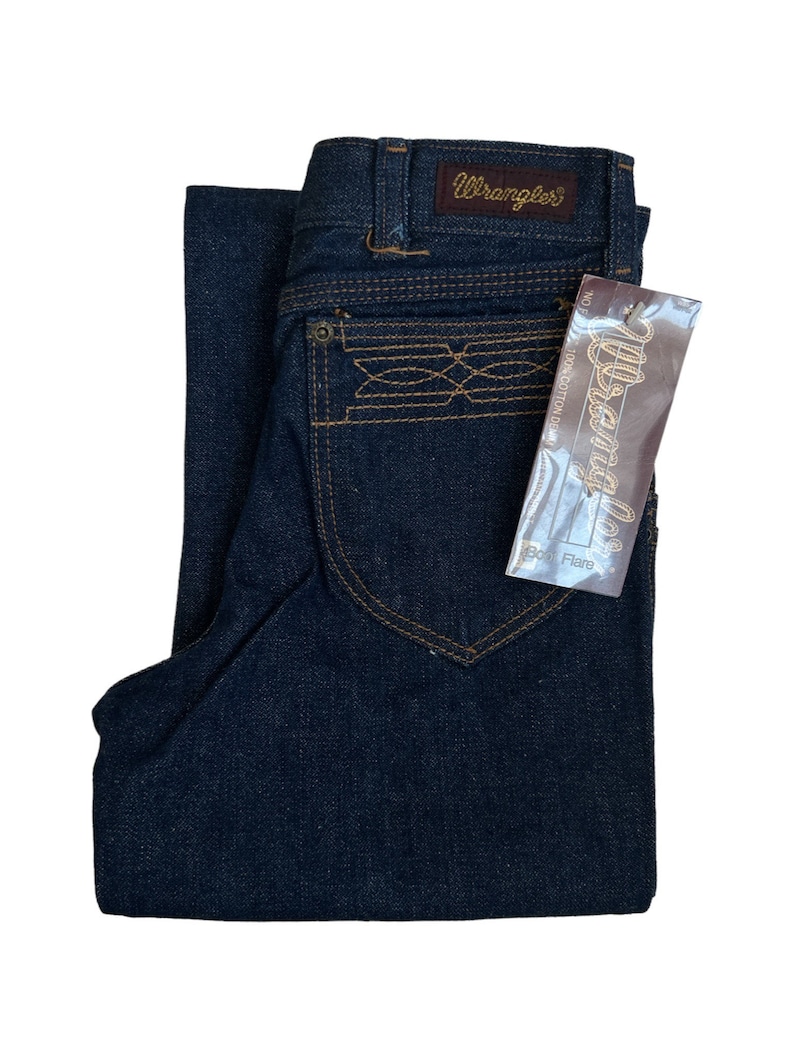 May include: A pair of dark blue denim jeans with a brown leather Wrangler label on the back waistband. The jeans have a boot cut flare and a brown embroidered design on the back pocket. The jeans have a tag that says "Wrangler" and "Boot Flare".