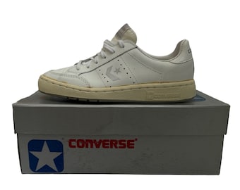 May include: White leather Converse sneakers with a white sole and a white star logo on the side. The shoes are in a white box with the Converse logo on the front.