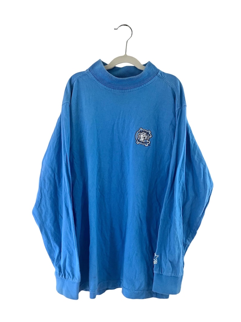 May include: A light blue long-sleeved t-shirt with a small embroidered logo on the front. The logo is a circle with a star inside and the words "Original Vintage" around it.