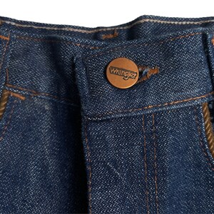 May include: Close-up of a pair of blue denim jeans with a brown leather patch that says "Wrangler" on the back pocket.