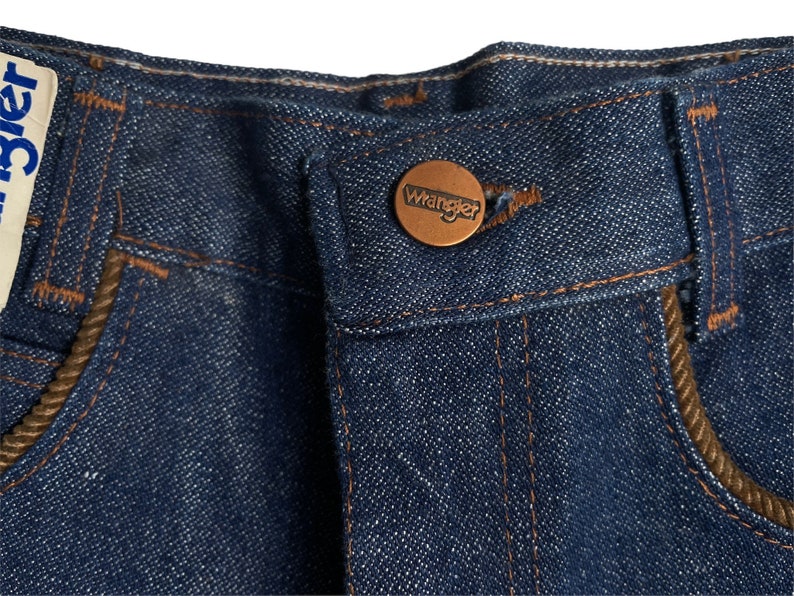 May include: Close-up of a pair of blue denim jeans with a brown leather patch that says "Wrangler" on the back pocket.