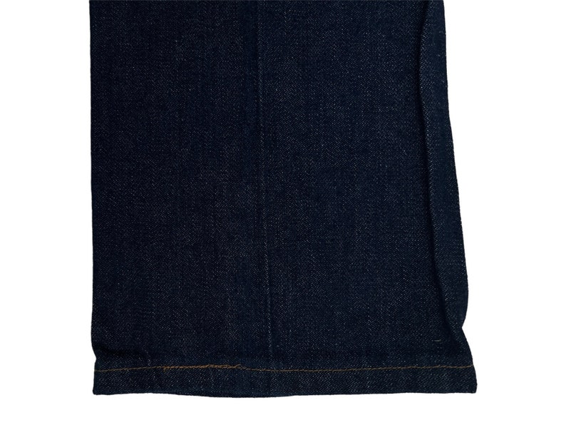 May include: Close-up of the bottom hem of a pair of dark blue denim jeans. The hem is sewn with a straight stitch and has a subtle brown thread.