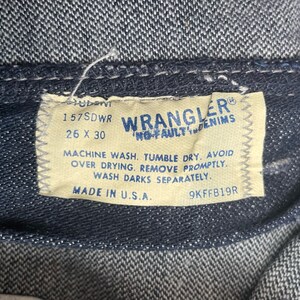 May include: A tan Wrangler denim label with the text "Wrangler No-Fault Denim" and the size "26 x 30". The label also includes the text "157SDWR", "Machine wash. Tumble dry. Avoid over drying. Remove promptly. Wash darks separately." and "Made in U.S.A. 9KFFB19R".