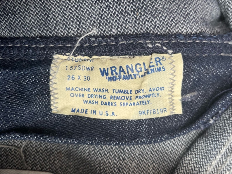 May include: A tan Wrangler denim label with the text "Wrangler No-Fault Denim" and the size "26 x 30". The label also includes the text "157SDWR", "Machine wash. Tumble dry. Avoid over drying. Remove promptly. Wash darks separately." and "Made in U.S.A. 9KFFB19R".