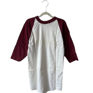 May include: A white and maroon baseball tee shirt on a hanger. The shirt has a white body and maroon sleeves.