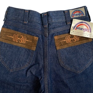 May include: A pair of blue denim jeans with brown corduroy pockets. The pockets have an embroidered design. The jeans have a Wrangler label on the back pocket and a "No-Fault!" tag on the right side.