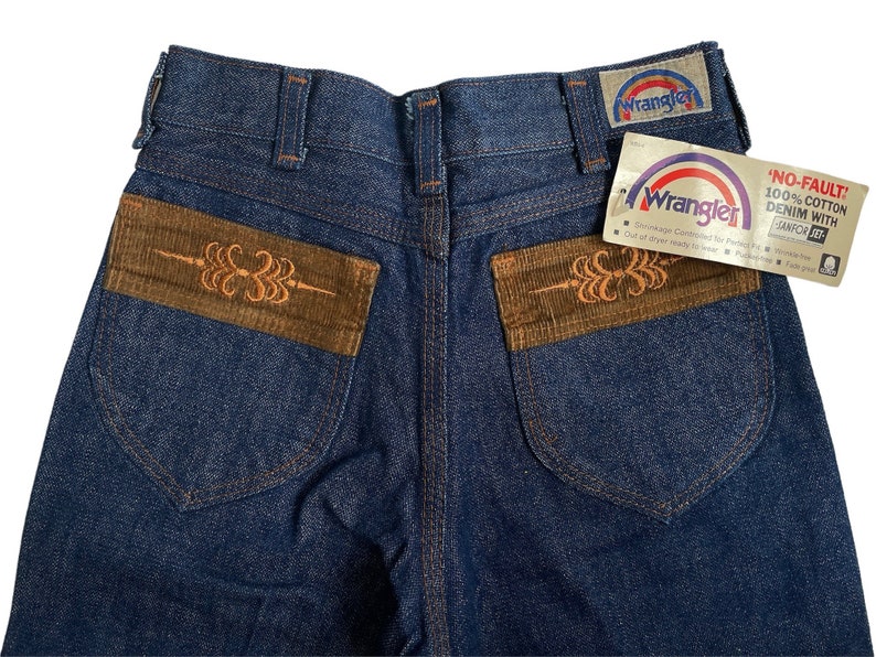 May include: A pair of blue denim jeans with brown corduroy pockets. The pockets have an embroidered design. The jeans have a Wrangler label on the back pocket and a "No-Fault!" tag on the right side.