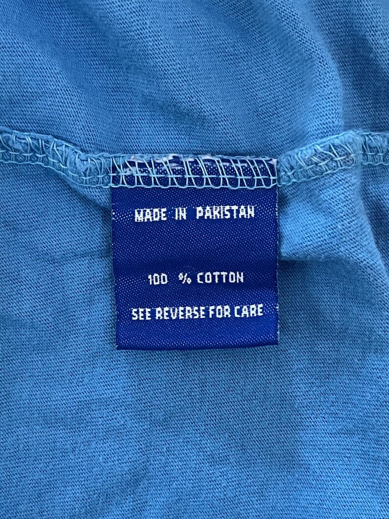 May include: Blue care label with white text that reads "Made in Pakistan", "100% Cotton", and "See Reverse for Care".