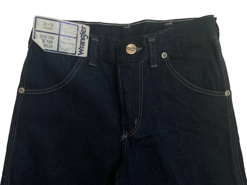 May include: A pair of dark blue denim jeans with a silver stud out flare, a Wrangler label, and a size tag that reads "26 x 30 Student".