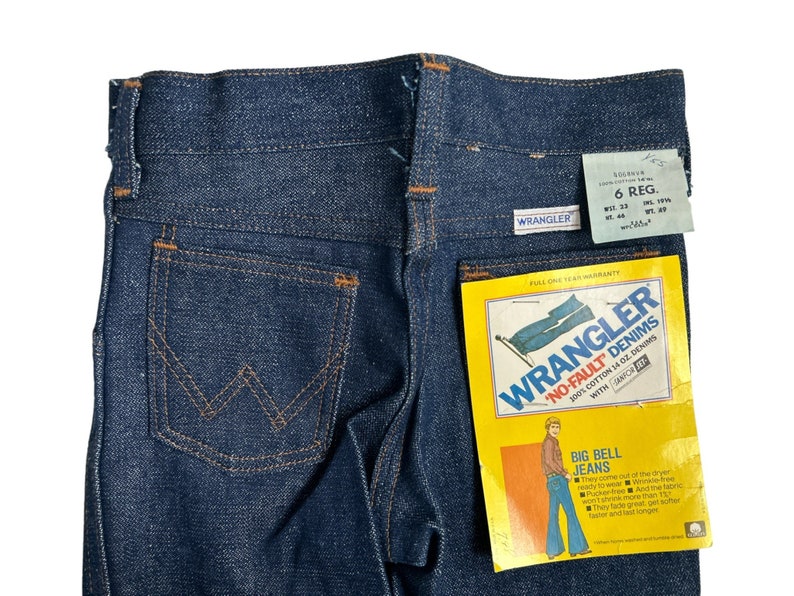 May include: A pair of blue denim jeans with a back pocket featuring the Wrangler logo. The jeans have a label that reads "Wrangler No-Fault Denims 100% Cotton 14 oz. Denims with Sanforized Fit" and "Big Bell Jeans".