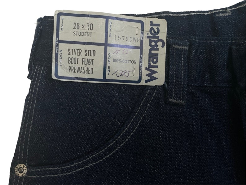 May include: A dark blue denim jean with a silver stud detail. The label reads "26 x 30 Student Silver Stud Boot Flare Prewasjed" and "Wrangler".