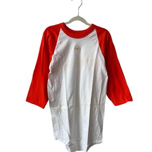 May include: White and orange baseball t-shirt with a size tag on the front. The shirt is hanging on a silver hanger.