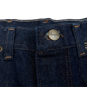 May include: Close-up of a pair of dark blue denim jeans with a brown leather Wrangler label sewn onto the waistband.