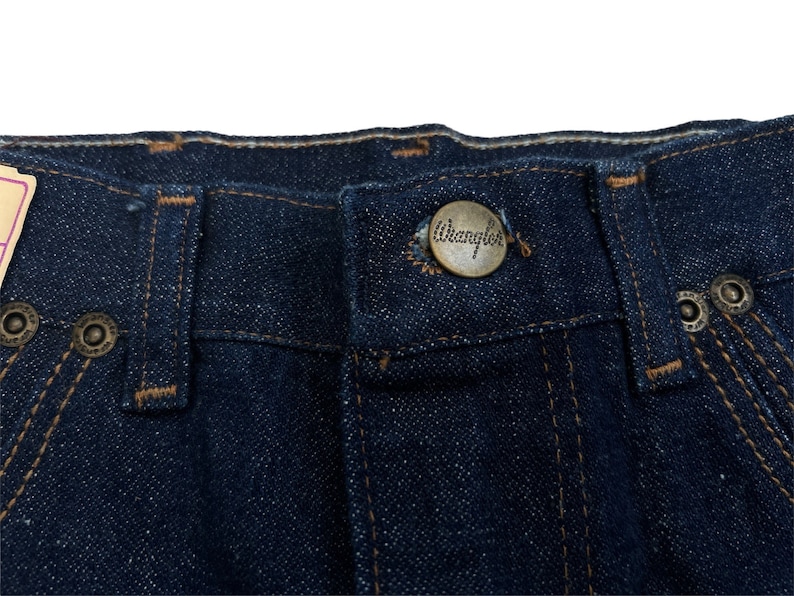 May include: Close-up of a pair of dark blue denim jeans with a brown leather Wrangler label sewn onto the waistband.
