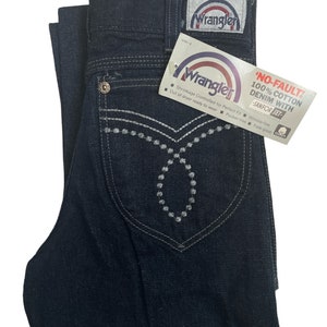 May include: A pair of dark blue denim jeans with a white embroidered design on the front pockets. The jeans have a Wrangler label on the back pocket and a tag that reads "No-Fault 100% Cotton Denim with Sanfor-Set".