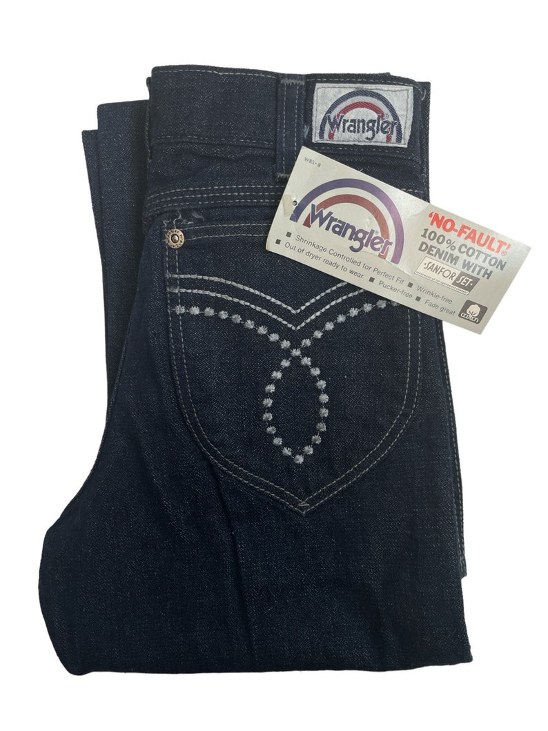 May include: A pair of dark blue denim jeans with a white embroidered design on the front pockets. The jeans have a Wrangler label on the back pocket and a tag that reads "No-Fault 100% Cotton Denim with Sanfor-Set".