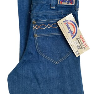 Może przedstawiać: Dżinsy z niebieskiego denimu z haftowanym wzorem w kolorze brązowym i białym na tylnej kieszeni. Dżinsy mają metkę Wrangler z logo tęczy.