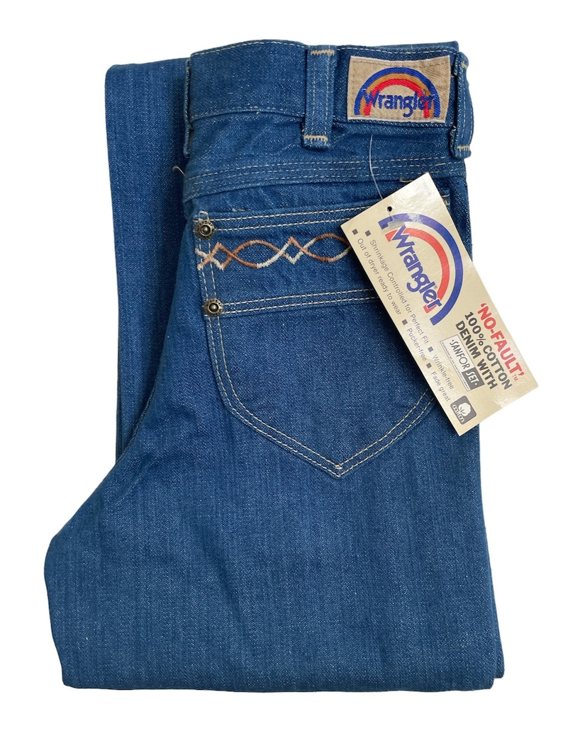 Może przedstawiać: Dżinsy z niebieskiego denimu z haftowanym wzorem w kolorze brązowym i białym na tylnej kieszeni. Dżinsy mają metkę Wrangler z logo tęczy.