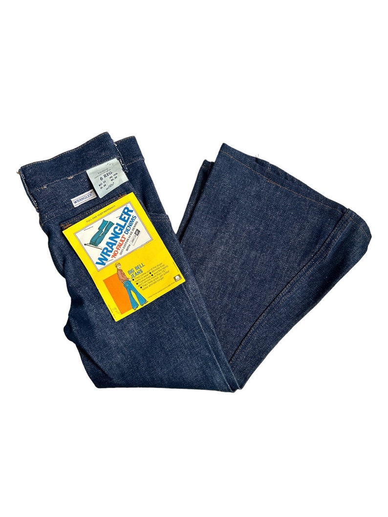 May include: A pair of dark blue denim bell bottom jeans with a yellow and orange Wrangler Big Bell Jeans tag attached. The tag features a cartoon illustration of a person wearing the jeans.