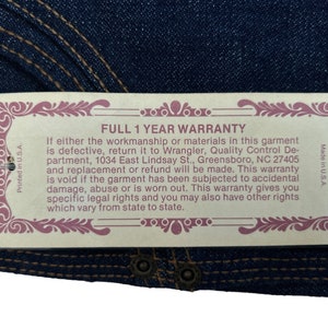 May include: A tan tag with a pink border and ornate design. The tag reads "FULL 1 YEAR WARRANTY" and includes details about the warranty, including the address of Wrangler's Quality Control Department in Greensboro, North Carolina.
