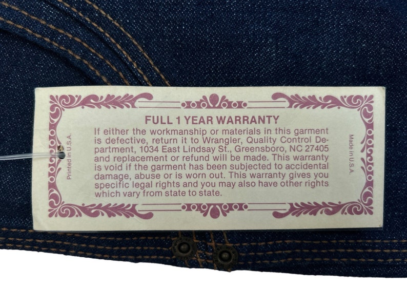 May include: A tan tag with a pink border and ornate design. The tag reads "FULL 1 YEAR WARRANTY" and includes details about the warranty, including the address of Wrangler's Quality Control Department in Greensboro, North Carolina.