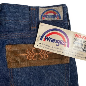 May include: A pair of blue denim jeans with a brown corduroy patch on the back pocket. The patch has an embroidered design. The jeans have a Wrangler label with a rainbow arc and the text 'NO-FAULT! 100% COTTON DENIM WITH SANFOR-SET' on the tag.