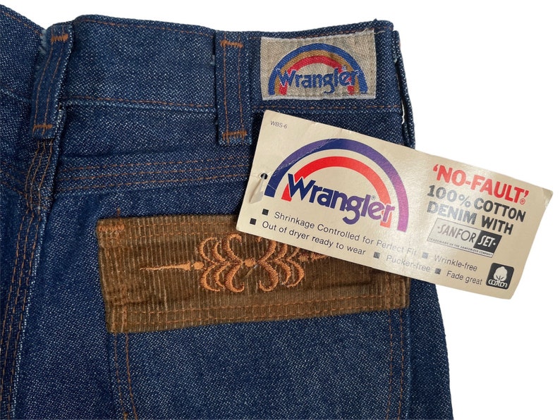 May include: A pair of blue denim jeans with a brown corduroy patch on the back pocket. The patch has an embroidered design. The jeans have a Wrangler label with a rainbow arc and the text 'NO-FAULT! 100% COTTON DENIM WITH SANFOR-SET' on the tag.