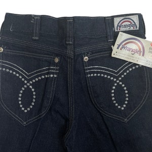 May include: Dark blue denim jeans with white stitching and a Wrangler label. The back pockets have a unique design with white stitching and small white dots.