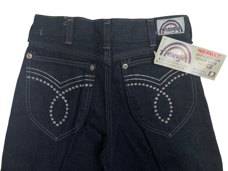 May include: Dark blue denim jeans with white stitching and a Wrangler label. The back pockets have a unique design with white stitching and small white dots.