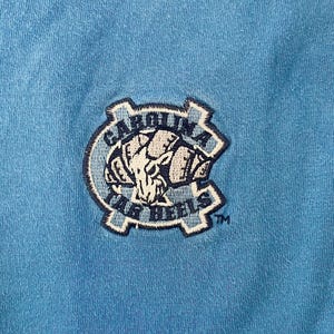 May include: A blue t-shirt with a navy blue embroidered patch featuring a ram's head and the text "Carolina Tar Heels" with a trademark symbol.