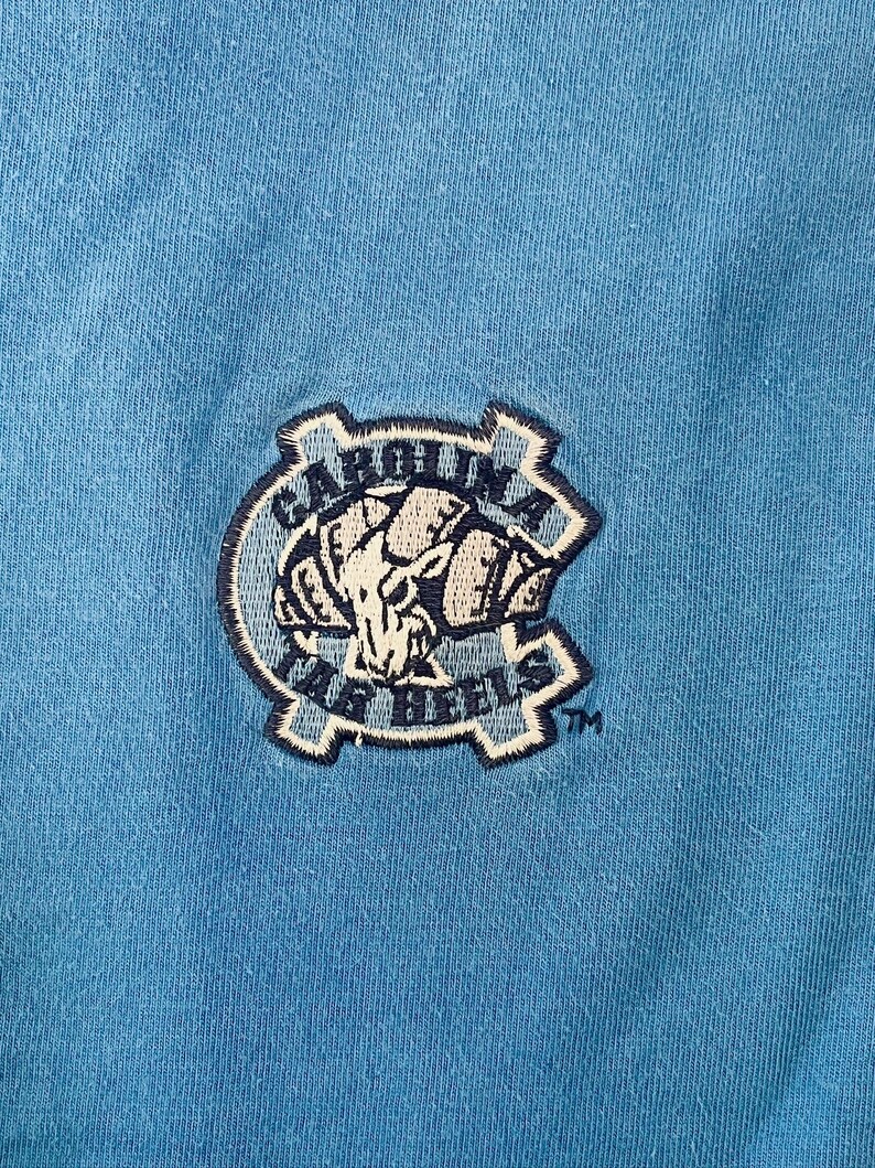 May include: A blue t-shirt with a navy blue embroidered patch featuring a ram's head and the text "Carolina Tar Heels" with a trademark symbol.