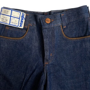 May include: A pair of blue denim jeans with brown stitching around the pockets. The jeans have a label that reads "Wrangler Lancer Boot Flare Prewashed 100% Cotton 7/8".