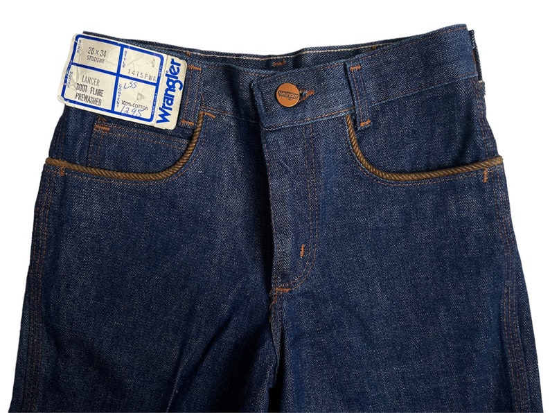 May include: A pair of blue denim jeans with brown stitching around the pockets. The jeans have a label that reads "Wrangler Lancer Boot Flare Prewashed 100% Cotton 7/8".