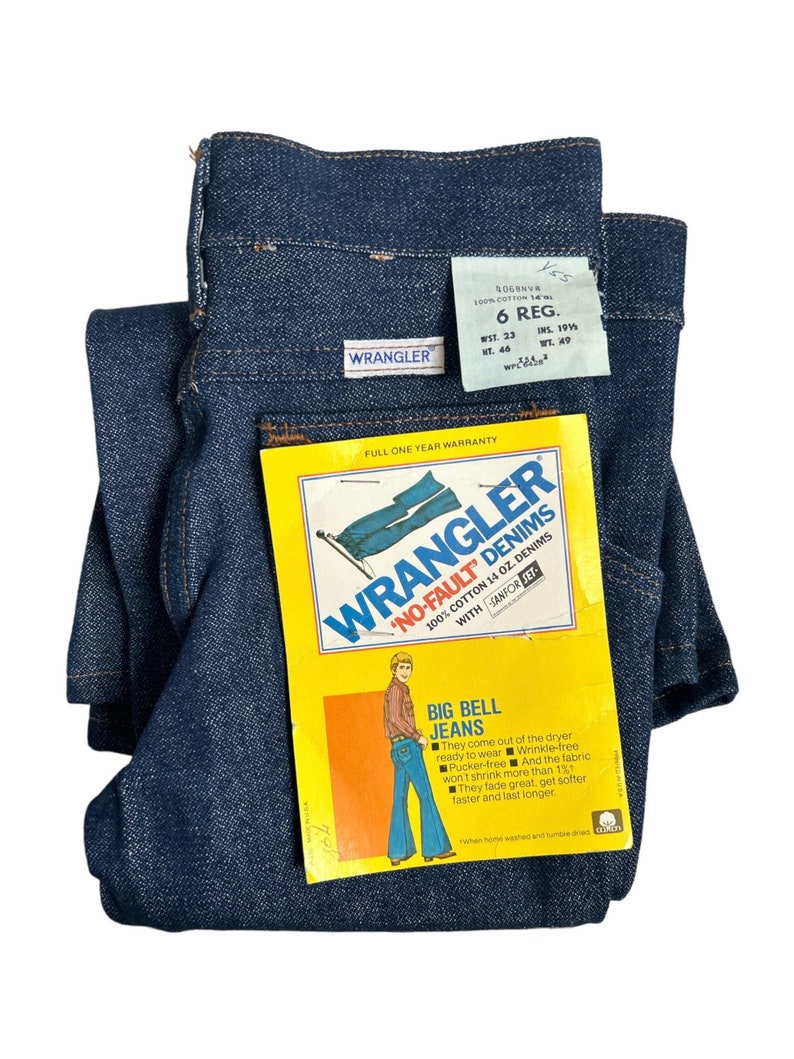 May include: A pair of blue denim Wrangler jeans with a tag attached. The tag is yellow and features a cartoon illustration of a person wearing flared jeans. The tag reads "Wrangler 'No-Fault' Denims 100% Cotton 14 oz. Denims with Sanfor-Set Big Bell Jeans".
