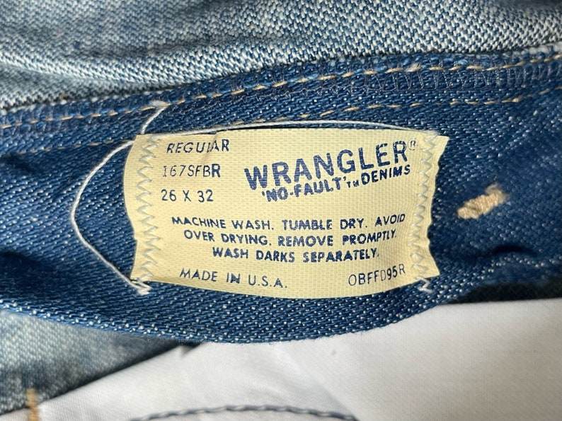 Może przedstawiać: Zbliżenie na niebieską metkę dżinsową z białym tekstem. Na metce widnieje napis "Regular 167SFBR 26 X 32 Wrangler 'No-Fault' Denim Machine Wash. Tumble Dry. Avoid Over Drying. Remove Promptly. Wash Darks Separately. Made in U.S.A. OBFFD95R"