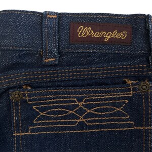 May include: Close-up of the back pocket of a pair of blue denim jeans. The pocket has a decorative stitching design and a small metal button. The jeans have a brown leather patch with the word "Wrangler" in gold lettering.
