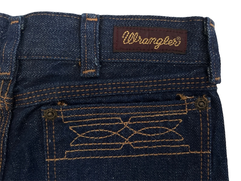 May include: Close-up of the back pocket of a pair of blue denim jeans. The pocket has a decorative stitching design and a small metal button. The jeans have a brown leather patch with the word "Wrangler" in gold lettering.