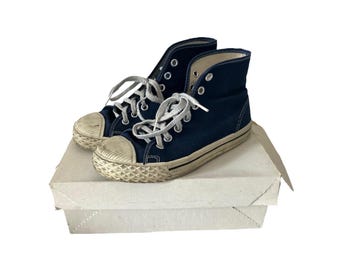 Puede incluir: Un par de zapatillas altas azul marino con cordones blancos y puntera de goma color crema. Los zapatos están colocados sobre una caja de cartón blanca, lo que sugiere un artículo vintage o de segunda mano.