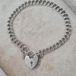 May include: A silver charm bracelet with a heart-shaped padlock clasp. The bracelet has a double-link chain design. The heart-shaped padlock features a keyhole and is attached to the chain with a small chain.