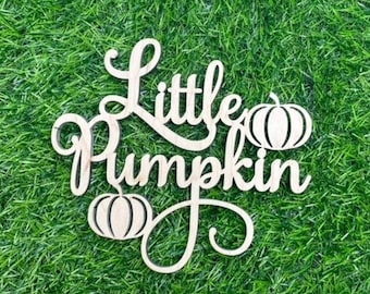May include: Wooden cutout of the words "Little Pumpkin" with a pumpkin design on either side of the words. The cutout is on a green grass background.