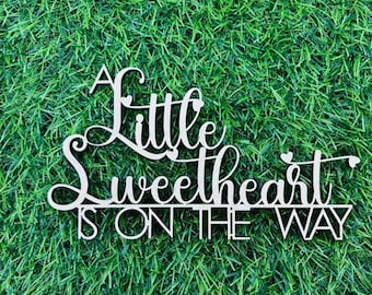 May include: White wooden sign with the text "A little sweetheart is on the way" with decorative hearts.