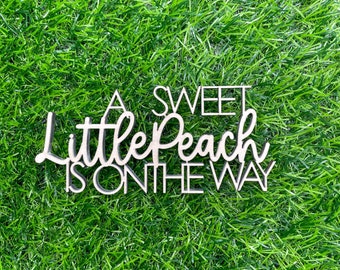May include: A wooden sign with the text "A Sweet Little Peach Is On The Way" in white lettering on a brown background.