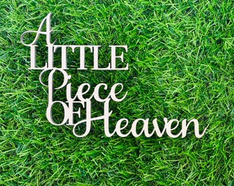 May include: A white wooden sign with the words "A Little Piece of Heaven" on a green grass background.