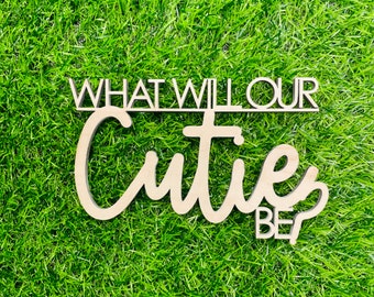 May include: Wooden sign with the text "What will our cutie be?" in a cursive font. The sign is on a green grass background.