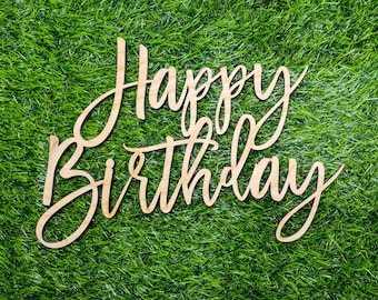 May include: Happy Birthday written in cursive on a wooden sign against a green grass background.