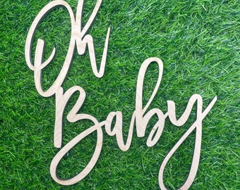 May include: Wooden sign with the words "Oh Baby" in a cursive font. The sign is made of light-colored wood and is set against a backdrop of green grass. The sign is a decorative item for a baby shower or nursery.