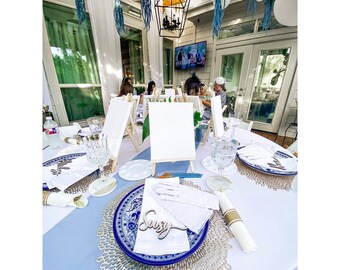 May include: A white table with blue and gold place settings. Each place setting has a white napkin, a blue and white plate, and a gold charger. There is a wooden name card with the name "Suzy" on it. There are also two white canvases on easels in the background.