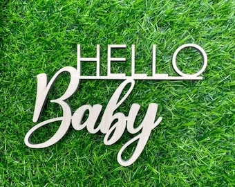 May include: Wooden sign with the words "Hello Baby" in white paint on a green grass background.