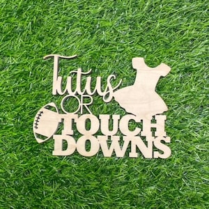 May include: Wooden sign with the text "Tutus or Touchdowns" in white lettering. The sign features a silhouette of a tutu and a football.